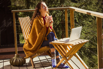 Relaxing woman on a wooden balcony enjoying coffee and working on laptop in the forest setting. A calm girl enjoys tea drink and sunlight on a wooden terrace, wrapped in a cozy blanket enjoying warmth
