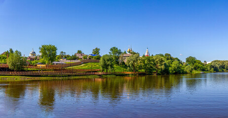 Serene panorama of Moskva River in Kolomna - rustic village houses and Orthodox church domes rise above trees, with wooden walkways on lush green bank. Perfect water reflections. Kolomna, Russia