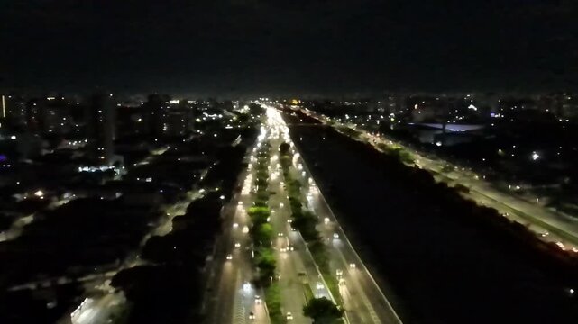 Aerial night view of the illuminated Marginal Tiet&ecirc; highway in S&atilde;o Paulo, Brazil, with heavy traffic flow, car light trails, and the dark Tiete River alongside the urban cityscape skyline.