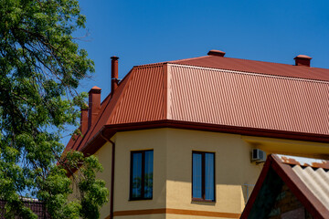 Modern house with distinctive reddish-brown corrugated metal roof and yellow walls against clear blue sky, partially framed by vibrant green tree foliage.
