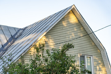 Residential house exterior featuring light-colored horizontal siding, corrugated metal roof and outdoor camera, illuminated by golden hour sunlight with dappled shadows and green foliage