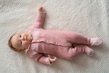Full-body portrait of cute baby wearing charming pink knitted sleepsuit with white accents, lying...