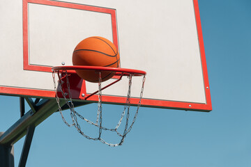 Naklejka premium Basketball orange ball in basketball hoop against clear blue sky. Game moment. Outdoors. Copy space.