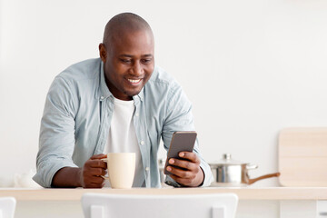 Happy african american man using smartphone and having morning coffee, standing in the kitchen, texting on cellphone, surfing internet and reading news, free space