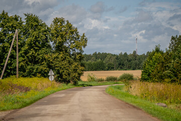 Curved asphalt country road through rural landscape with green trees, yellow fields, road signs and communication tower under a partly cloudy summer sky