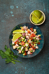 Fresh and vibrant peruvian seafood ceviche with shrimps, octopus, tomatoes, cucumber and parsley, served with lime in a bowl on a stone background from above, dish of Peru