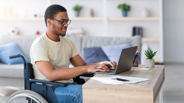 Black guy in wheelchair working online from home, using laptop, looking for job opportunities on internet. Young man with disability having business conference, making business project on web
