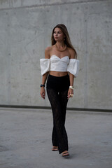 Fototapeta premium Stylish young woman in fashionable clothes posing confidently outdoors. Fashion model look with urban background, long hair, black shoes, and modern accessories.