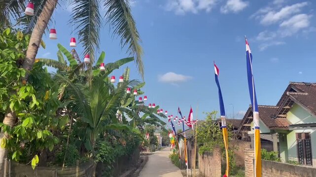 A rural Indonesian street decorated for Independence Day celebrations. Red and white flags, colorful bamboo poles (penjor), and festive ornaments line the path under a bright blue sky. Traditional hou