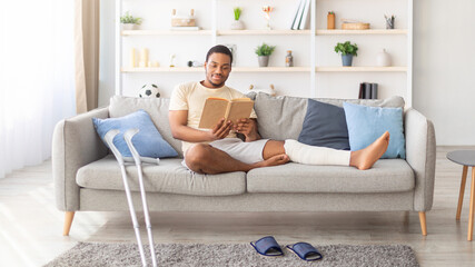 Millennial black guy with plastered leg sitting on sofa, reading book at home, pair of crutches standing nearby. Young African American man having broken limb, resting on couch with interesting story