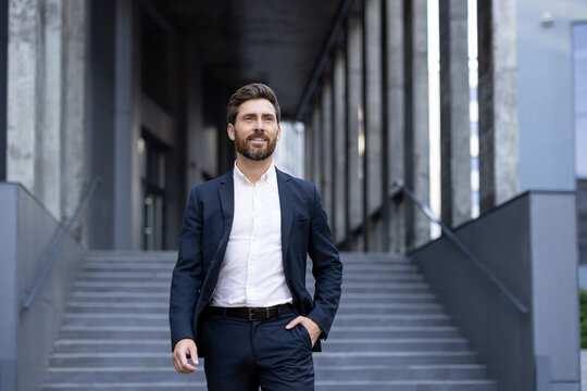 A confident businessman dressed in a formal suit walks past an urban office setting. The modern architecture and professional demeanor convey success, professionalism