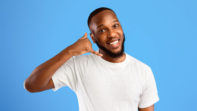 Flirty African Guy Showing Call Me Gesture Holding Hand Near Ear Like Telephone Standing Over Blue Studio Background, Smiling To Camera. Contact Me, Mobile Communication Concept. Panorama