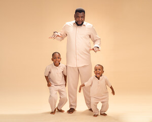 Smiling Black father posing barefoot with two young sons in matching beige outfits, joyful family portrait in studio with warm beige background, playful bonding and happiness together