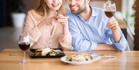 Affectionate millennial couple taking photo of yummy food and wine on smartphone at restaurant. Pair of sweethearts with mobile device celebrating Valentine's Day or anniversary together, cropped