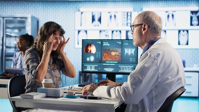 Senior woman at checkup deals with a painful headache and asks for advice at medical facility, recommending a treatment to relieve pain and ease the inflammation. Health insurance. Camera A.