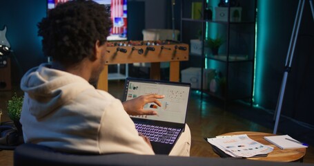 African american person working from home and checking statistical data on laptop screen, analyzing financial graphs. Teleworker in the living room comparing business revenue numbers. Camera B.