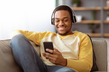 Joyful african american young man watching videos on mobile phone while sitting on couch at home, using wireless headset, copy space. Smiling black guy watching movie on smartphone