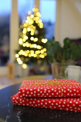 Two beautifully wrapped red gifts with white polka dots are placed on a table, with a softly lit Christmas tree twinkling in the background, creating a warm holiday atmosphere.