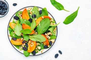 Autumn vegan persimmon salad with walnuts, blue raisin and spinach, white background, top view