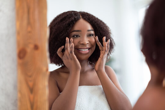 Charming black lady smiling at herself in mirror, checking first wrinkles, admiring soft silky skin after face cream at home. African American woman looking at her reflection after skincare treatment - Powered by Adobe