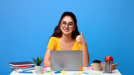 Portrait of positive smiling indian woman in eyeglasses showing thumbs up gesture sitting at the...