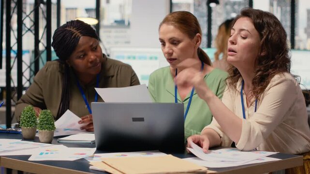 Businesswomen compare analytics and plan strategy in a modern workspace, supporting development vision together. Diverse associates collaborating and reviewing data analytics. Camera B.