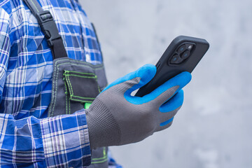 Close-up of a construction worker handyman in protective gloves and workwear using a modern smartphone. Concept digital technology into manual labor and job.
