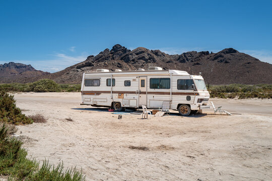 An old Class A motorhome with chairs parked on a Baja Mexico beach with desert mountains in the background.