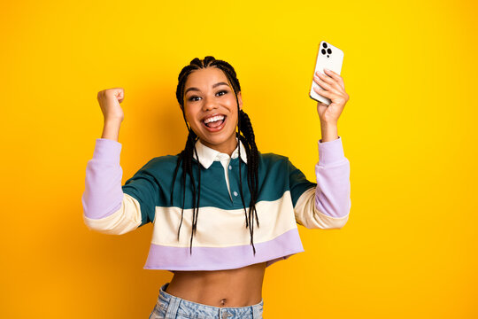 Joyful young woman with braided hair celebrating while holding phone against bright yellow background