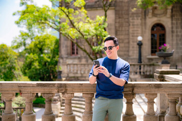 Fototapeta premium Happy young Latino man with short hair, wearing sunglasses, navy shirt and green pants, stands at Chapultepec Castle in Mexico City smiling while checking his phone in a relaxed, happy mood