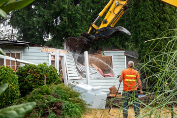 Workman with hose spraying water on demolition of old wood frame house for redevelopment of...