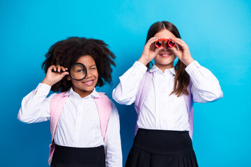 Cheerful schoolgirls using magnifying glass and binoculars with playful expressions against a vibrant blue background
