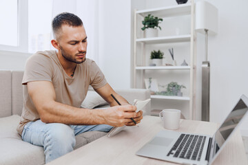 Thoughtful man taking notes at home office, dressed casually, engaged in creative process with a laptop nearby, plants in background, modern aesthetic.
