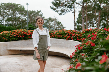 Young woman smiling in a garden with flowers, wearing stylish green overalls and a white shirt, conveying happiness in a bright outdoor setting