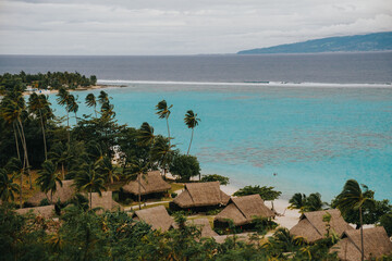 Thatched beach bungalows line a tropical shoreline with turquoise waters on Moorea Island, French Polynesia.