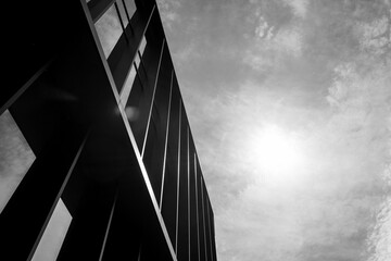 Black and white photo of a modern building facade with strong vertical lines and sunlight in a cloudy sky. Suitable for illustrating architecture, urban design, or minimalism concepts in editorial and