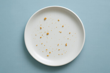 Empty white plate with scattered cookie crumbs on blue background, minimalistic top view symbolizing finished meal or snack