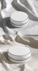 Two identical white, round, compact devices displayed on draped fabric.  Clean, minimalist design.  Soft lighting accentuates the form
