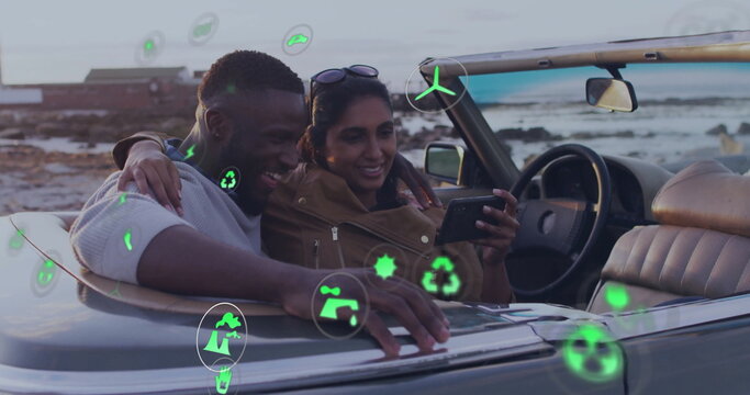 Leaning couple with sunglasses viewing smartphone in convertible by coast with floating green icons