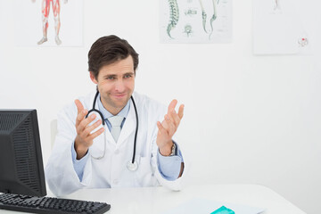 Mid adult male physician in lab coat sitting in clinic with stethoscope, explaining anatomy charts