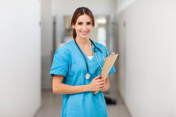 Female nurse standing in hospital corridor in blue scrubs and stethoscope holding clipboard smiling