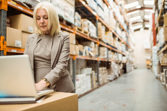 Senior woman wearing business suit working on laptop atop box in warehouse aisle racks, copy space