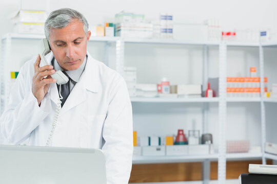 Male pharmacist talking on corded phone checking laptop at pharmacy counter, copy space