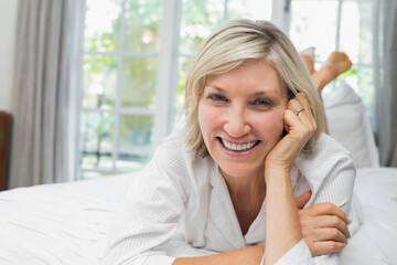 Senior woman wearing pajamas lying on white sheets in bedroom smiling at camera by sheer curtains