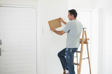 Wooden picture frame being leveled on white hallway wall using stepladder near shuttered window