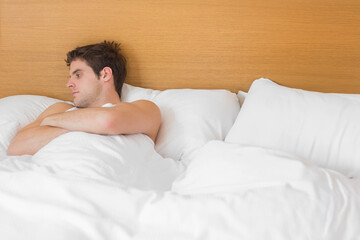 Man reclining against wooden headboard on bed wearing sleeveless undershirt under duvet, copy space