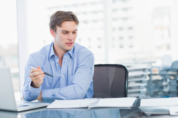 Fototapeta premium Male business professional reviewing documents at glass desk in modern office with laptop and pen