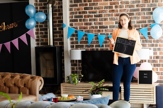 Female holding chalkboard tallying votes for gender reveal in living room with balloons, copy space