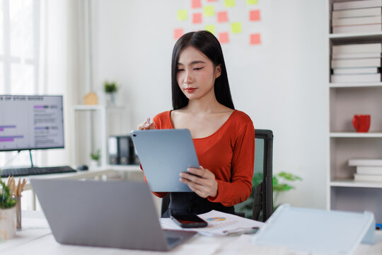 Asian Female business person using digital tablet and laptop or remote working at office, looking at digital tablet learning training, studying online thinking Financial and Accounting concept.
