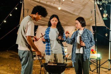 Group of Asian friends enjoying a camping barbecue at night under warm firelight. Woman grills colorful skewers next to warm tent, creating a fun and friendly outdoor dining atmosphere.